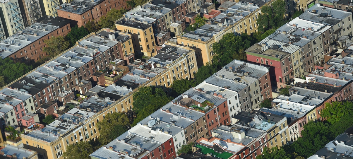 Aerial view of several city blocks with rows of apartment buildings featuring white roofs, separated by tree-lined streets in an urban neighborhood. Aerial view of several city blocks with rows of apartment buildings featuring white roofs, separated by tree-lined streets in an urban neighborhood.
