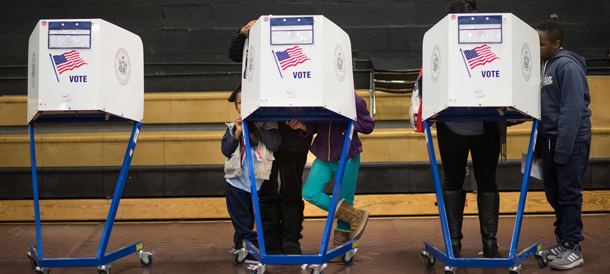 Three voting booths with “VOTE” and an American flag are occupied by people casting ballots in a gymnasium. Some children are visible, including one peeking from under a booth. Wooden bleachers are in the background. Three voting booths with “VOTE” and an American flag are occupied by people casting ballots in a gymnasium. Some children are visible, including one peeking from under a booth. Wooden bleachers are in the background.