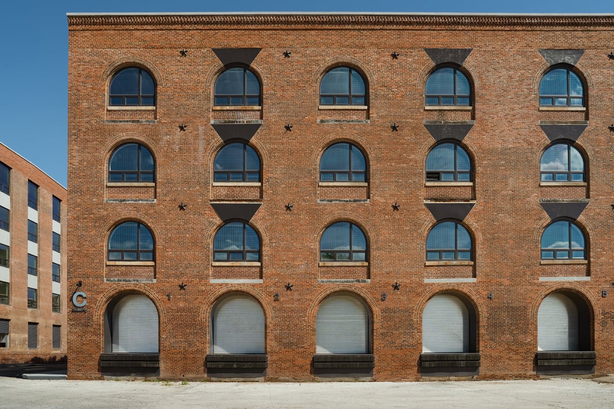A brick building with three floors and three arched windows on each level, each window framed with dark accents. The ground floor windows appear to have closed metal shutters. The sky is clear and blue.
