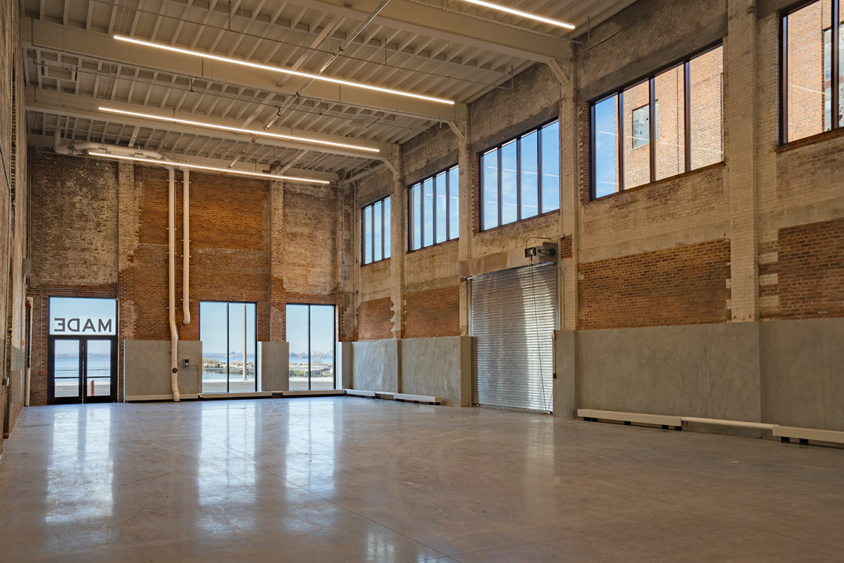 Spacious industrial warehouse with high ceilings, exposed brick walls, large windows letting in natural light, and a rolled-up metal garage door; outside, water and buildings are visible through the windows and glass doors.