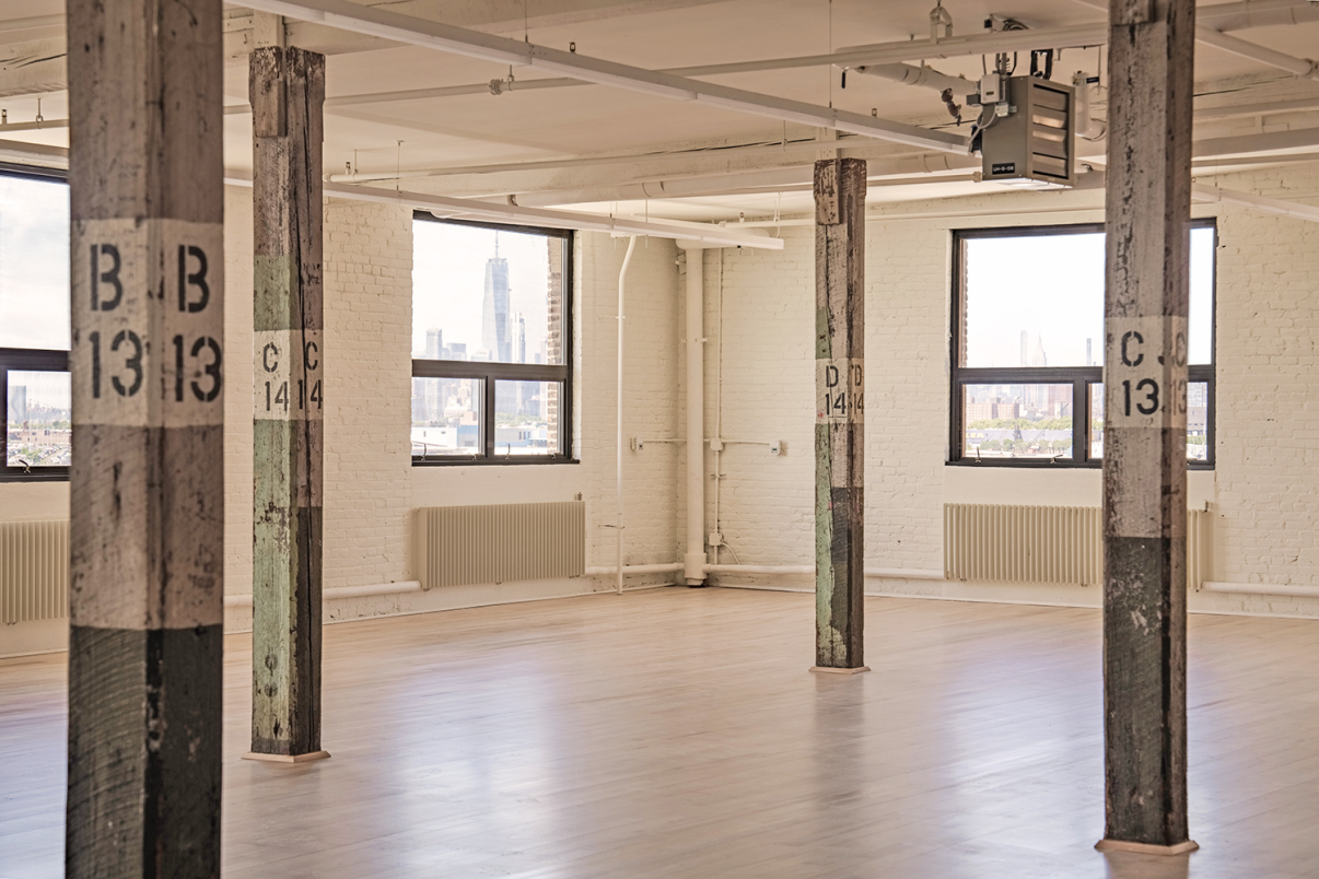 Spacious industrial loft with wooden pillars labeled B13 and C14, large windows offering city skyline views, and light wood flooring. Walls are white brick with a clean, minimalist appearance.