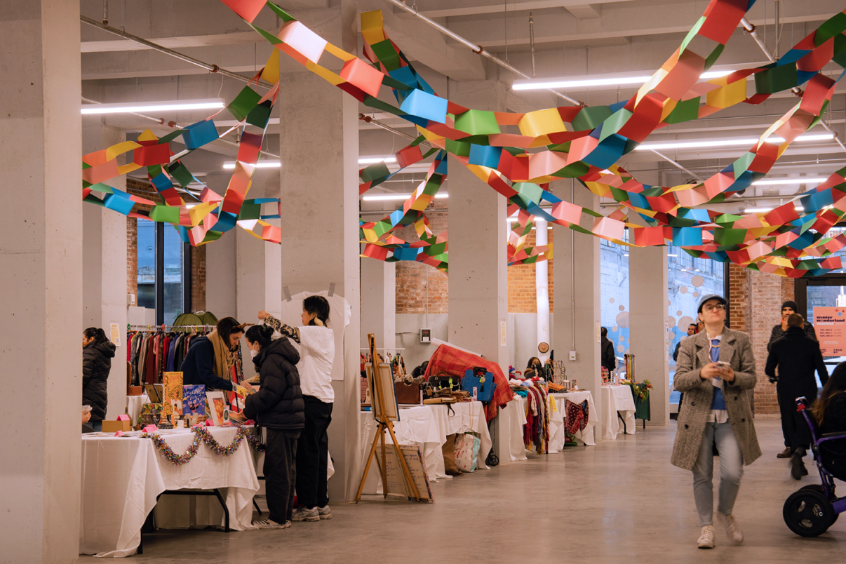 A spacious indoor market with colorful paper garlands hanging from the ceiling. Several people browse tables displaying various items like clothes, art, and crafts. A woman walks past with a stroller on the right.