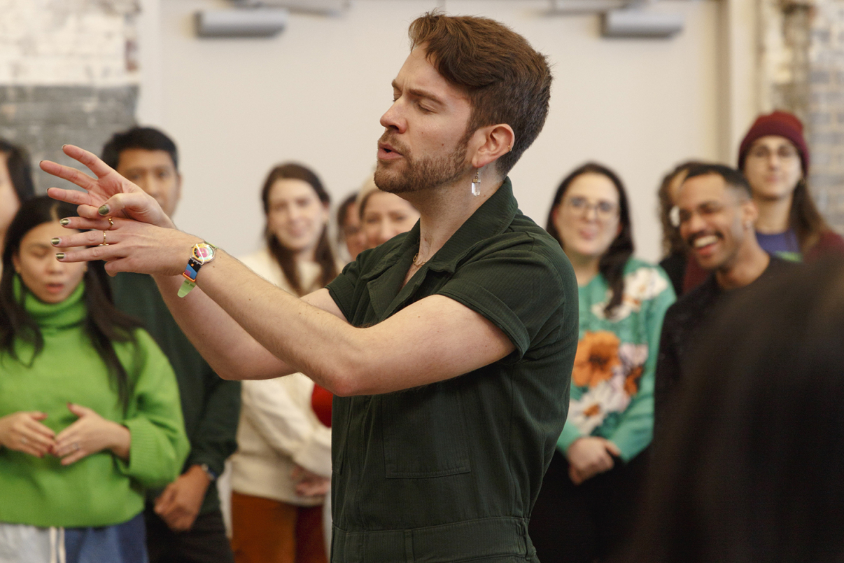 A person with painted nails and an earring gestures expressively while speaking or performing, surrounded by a diverse group of people who are watching and smiling in a casual indoor setting.