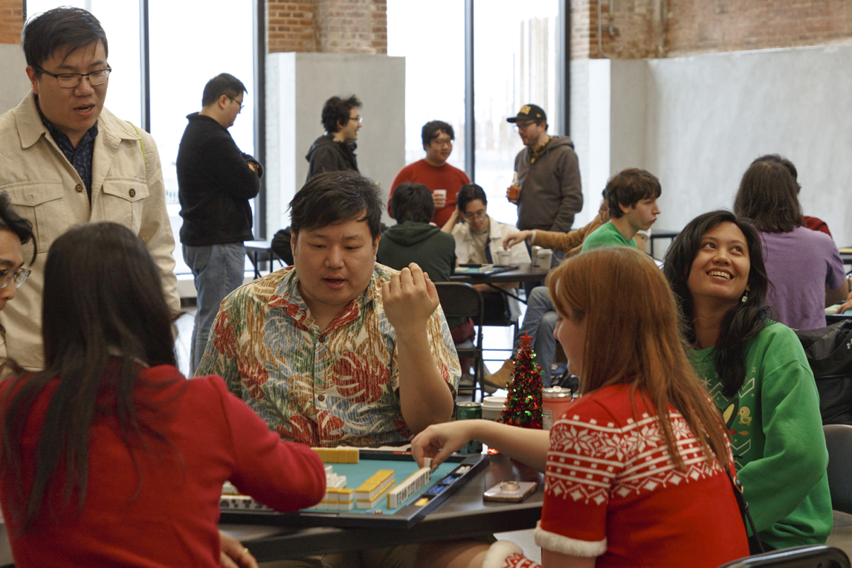 Show thumbnail preview A group of people sits around tables playing games indoors. In the foreground, four people play a board game while others socialize in the background. The atmosphere appears lively and friendly.