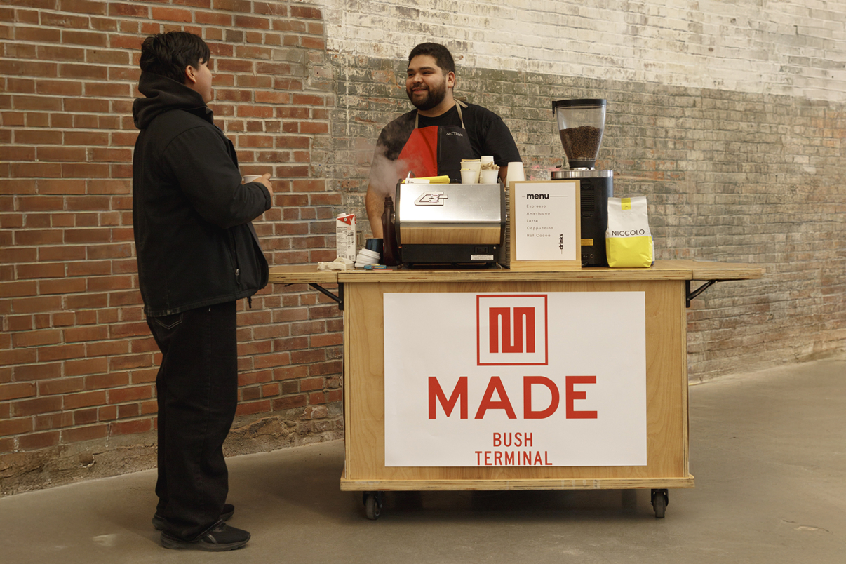 A barista smiles and talks with a customer at a small coffee cart labeled MADE BUSH TERMINAL, set against a brick wall. The cart holds a coffee machine, cups, and coffee supplies.