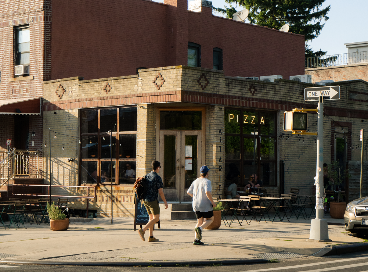 Two people walk past a brick corner pizza shop with large windows on a sunny day. The street is quiet, and there are plants and outdoor seating in front of the restaurant. A tree and a parked car are visible in the background.