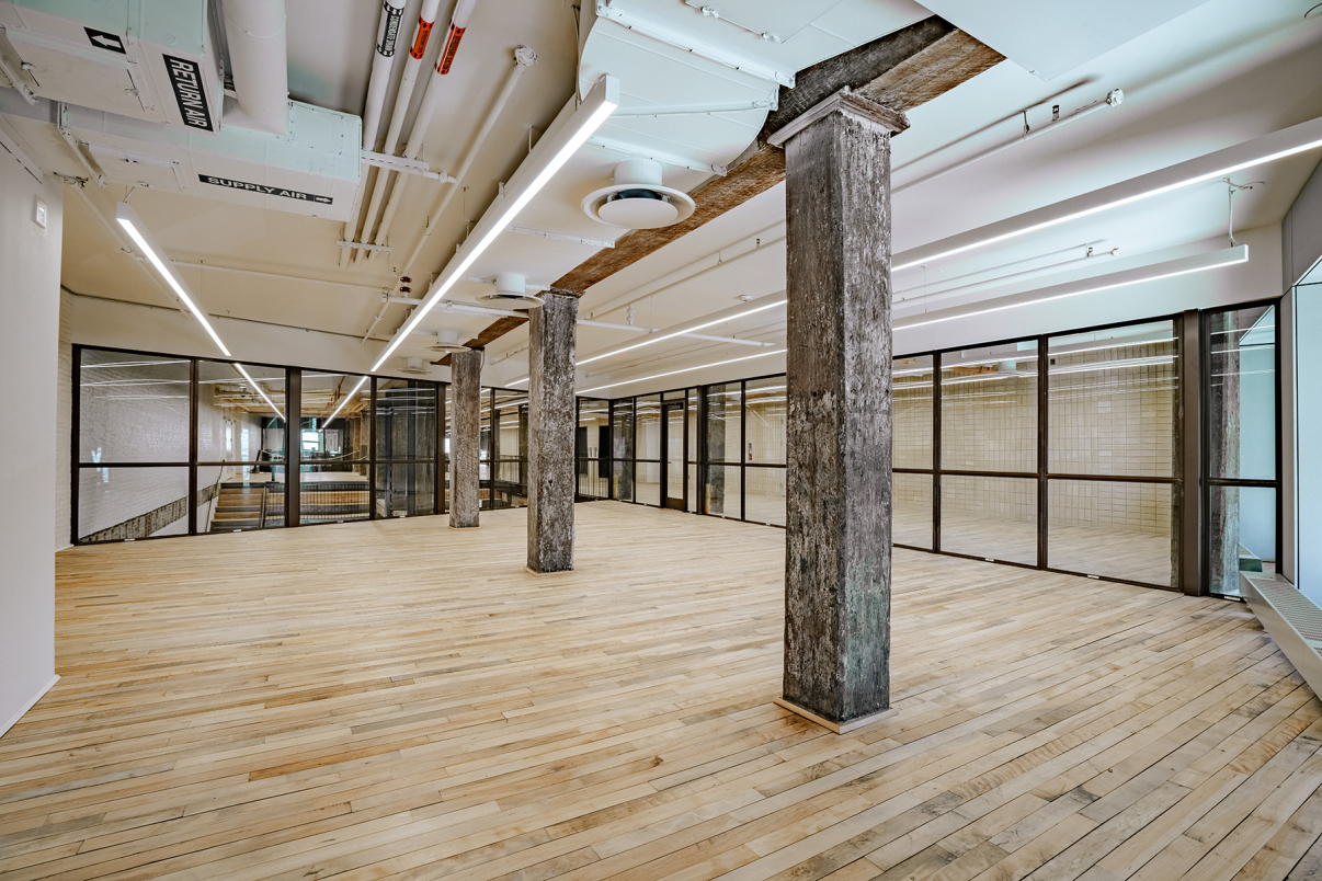 Modern open-plan office space with light wooden floors, exposed concrete columns, visible ceiling pipes, and glass walls dividing rooms. Bright fluorescent lights illuminate the spacious, empty interior.
