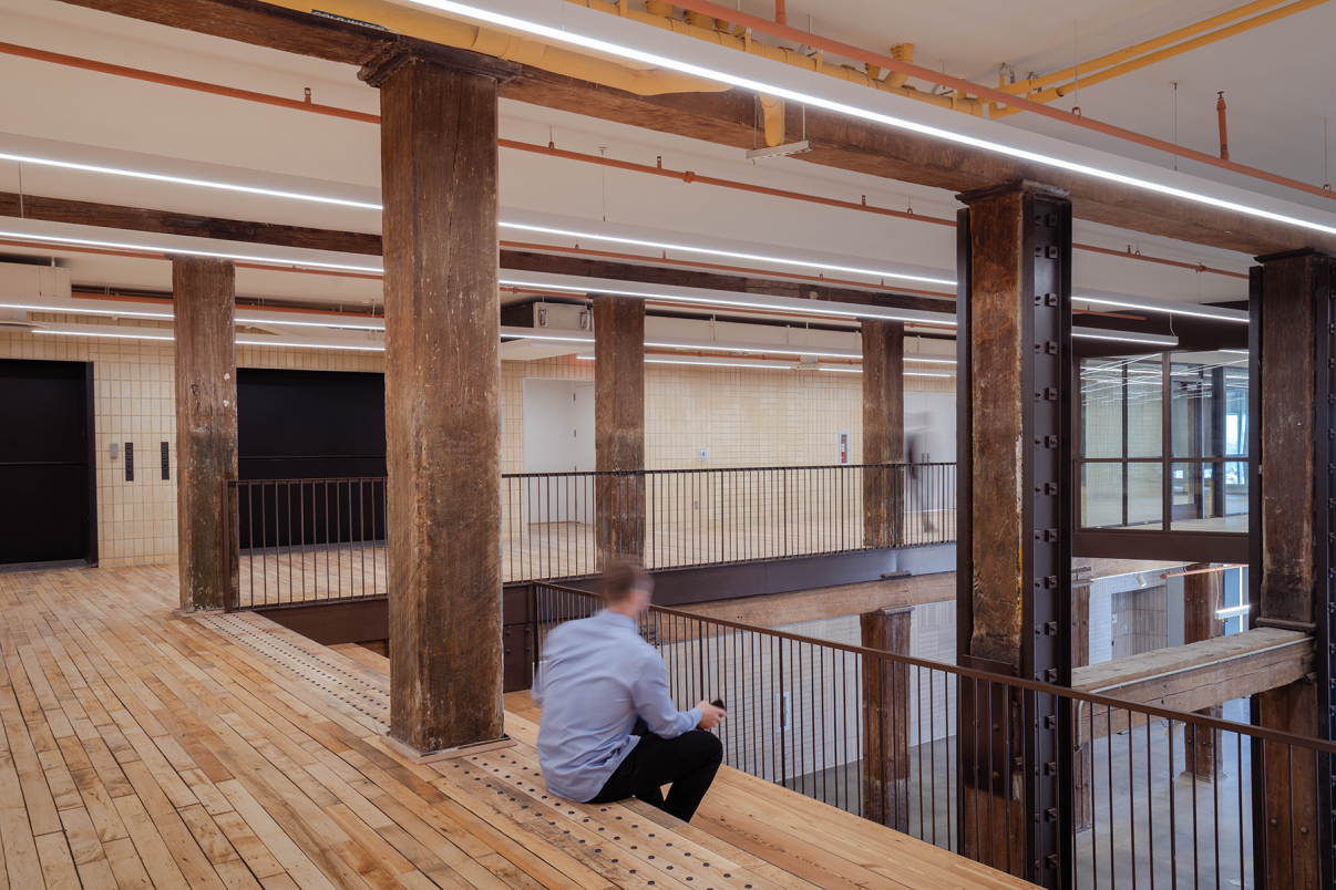 A person sits on a wooden bench in an industrial-style interior with exposed beams, wooden floors, metal railings, and cream tile walls, creating a spacious, modern atmosphere.