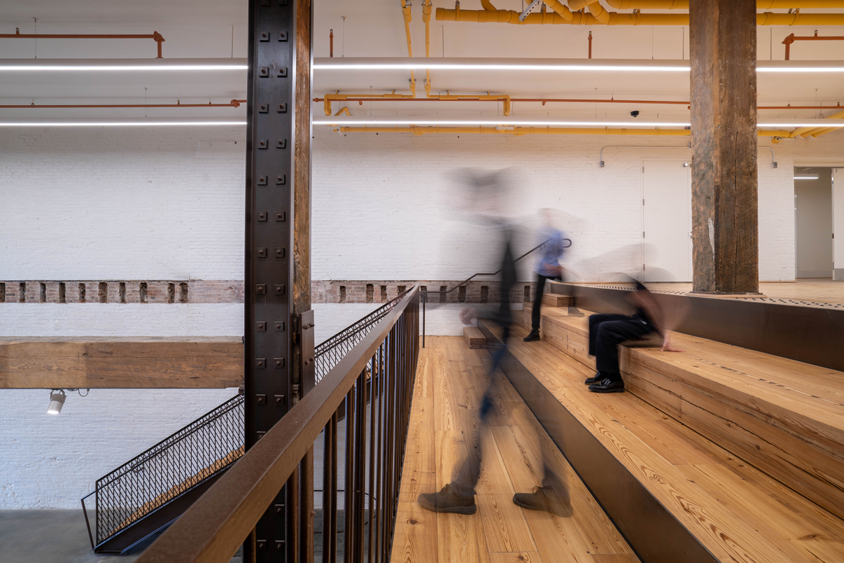 A modern interior with wooden stadium-style seating and exposed beams; three blurred figures move across and up the steps, suggesting motion. White brick walls and yellow overhead pipes are visible in the background.