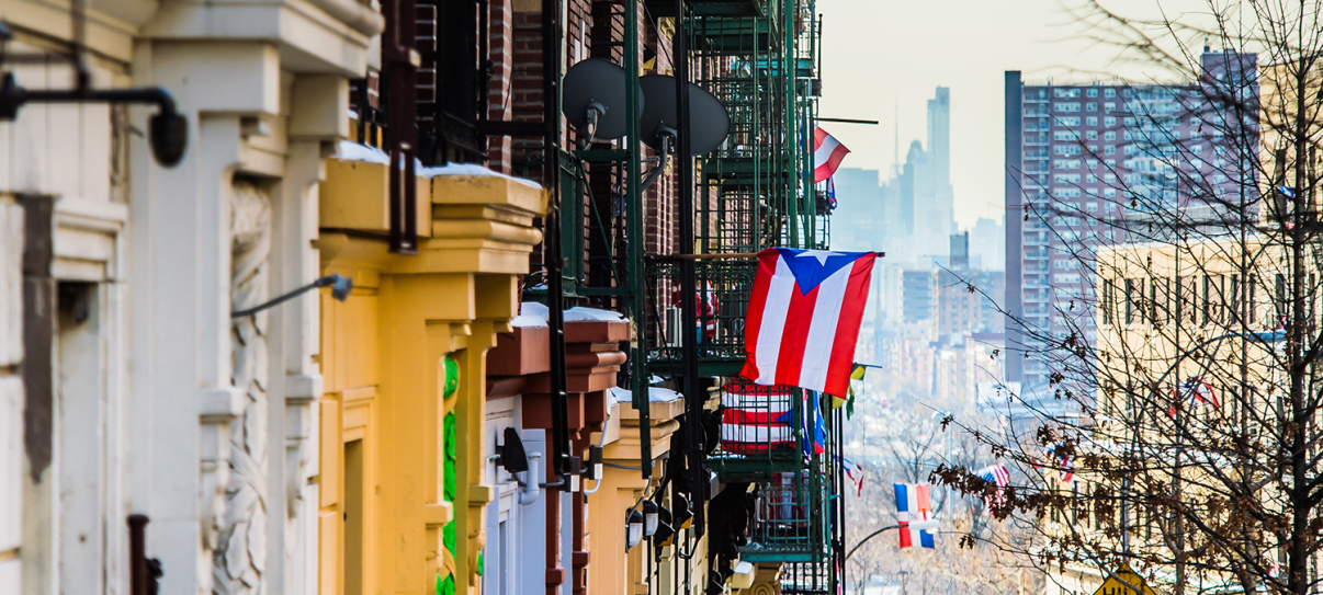 Colorful apartment buildings line a New York City street, with Puerto Rican flags hanging from fire escapes. In the background, high-rise buildings and a hazy skyline are visible. Leafless tree branches frame the right side of the image. Colorful apartment buildings line a New York City street, with Puerto Rican flags hanging from fire escapes. In the background, high-rise buildings and a hazy skyline are visible. Leafless tree branches frame the right side of the image.