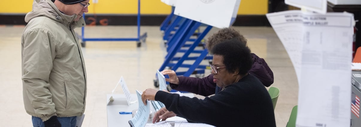 A poll worker checks a document while assisting a voter wearing a jacket and beanie at a polling station; voting booths are visible in the background. A poll worker checks a document while assisting a voter wearing a jacket and beanie at a polling station; voting booths are visible in the background.