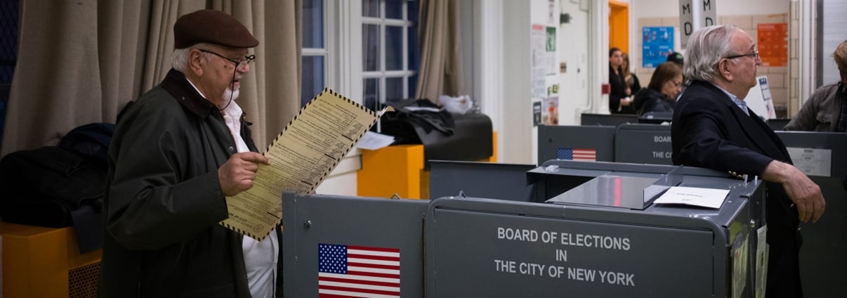 An elderly man holding a ballot stands at a voting booth in New York City. Another man sits at a table nearby. Voting booths and a Board of Elections in the City of New York sign are visible. An elderly man holding a ballot stands at a voting booth in New York City. Another man sits at a table nearby. Voting booths and a Board of Elections in the City of New York sign are visible.