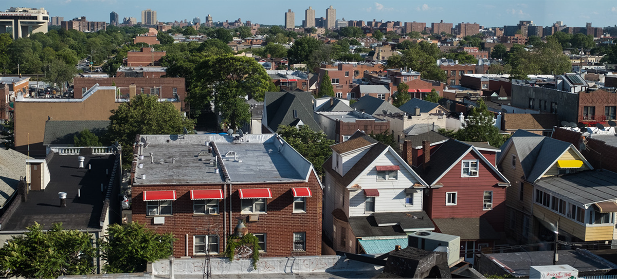 Show thumbnail preview A view of a city neighborhood with rows of houses, red brick buildings, green trees, and distant apartment towers under a blue sky with some clouds. Show thumbnail preview A view of a city neighborhood with rows of houses, red brick buildings, green trees, and distant apartment towers under a blue sky with some clouds.