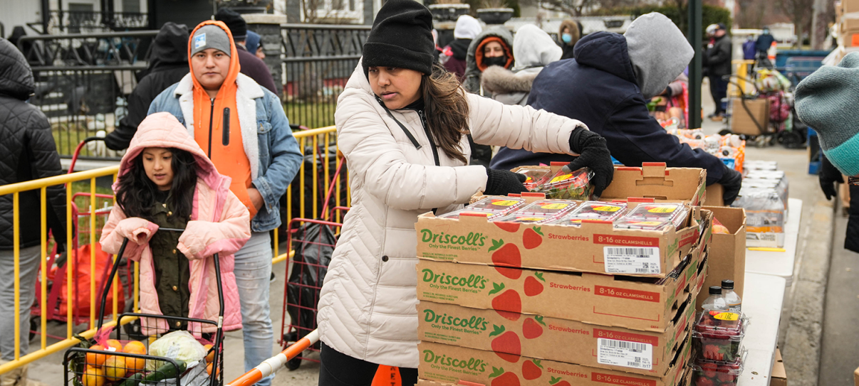 People bundled in warm clothing line up outdoors at a food distribution event, where a woman in a white jacket hands out boxes of strawberries beside stacked crates and shopping carts filled with groceries. People bundled in warm clothing line up outdoors at a food distribution event, where a woman in a white jacket hands out boxes of strawberries beside stacked crates and shopping carts filled with groceries.