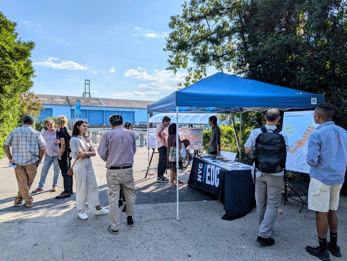 People gather at Brooklyn Bridge Park near a blue tent with an NYC/EDC table, engaging in conversation and viewing maps and posters under sunny skies, surrounded by greenery and industrial buildings.