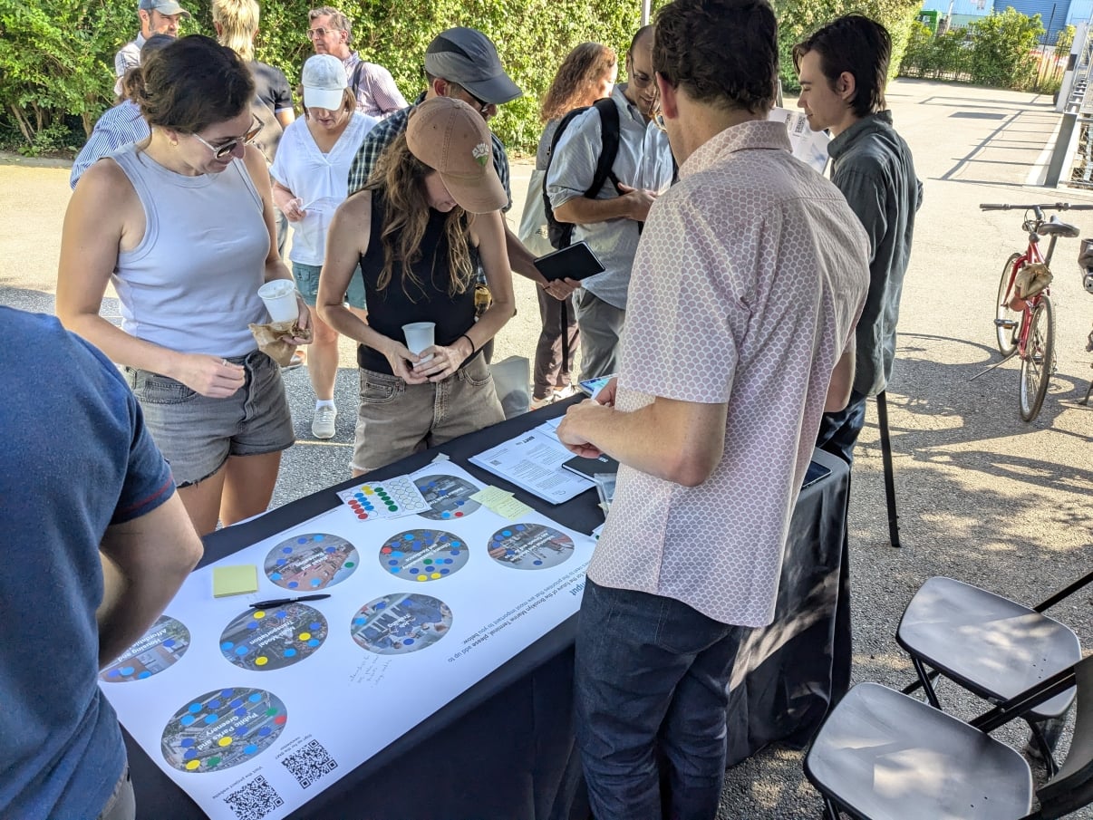 A group of people gathers around a table at Brooklyn Bridge Park for a Brooklyn Marine Terminal community event, examining maps and placing colored stickers. Some hold papers or drinks, and bicycles are visible in the background.