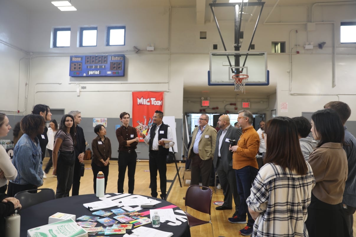 A group of adults stands in a circle in a gymnasium, listening to a person speaking, at the first Brooklyn Marine Terminal Public Workshop. Tables with papers and books are in the foreground, and a red banner and scoreboard are visible in the background.