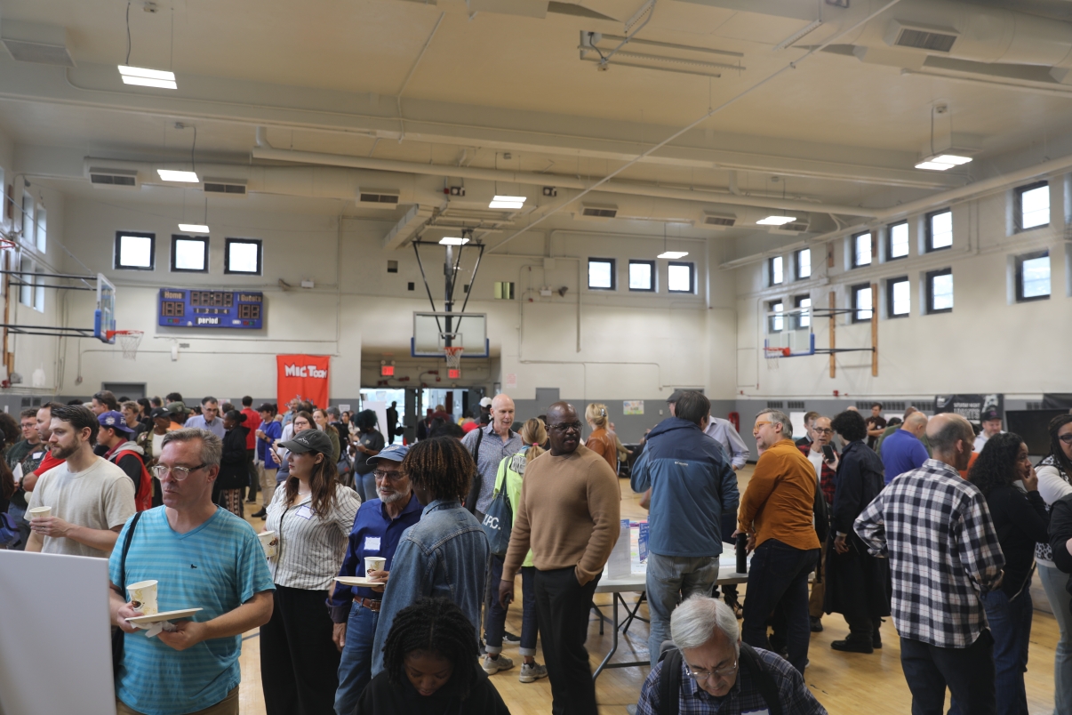Diverse group of people socialize and enjoy refreshments at the first Brooklyn Marine Terminal Public Workshop inside a gymnasium. Tables are set up, and some attendees hold cups and plates. Basketball hoops and scoreboard are visible in the background.