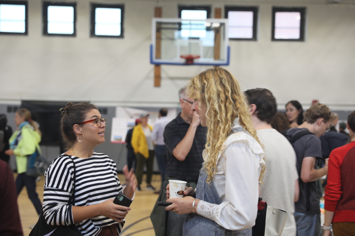 Two women stand in a gymnasium, engaged in conversation during the first Brooklyn Marine Terminal Public Workshop. One wears a striped shirt and glasses, the other has long curly hair and holds a cup. Other people and a basketball hoop are visible in the background.