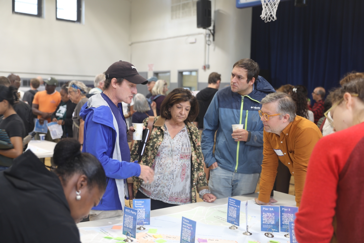 Group of people gather around a table with maps and informational signs at the first Brooklyn Marine Terminal Public Workshop in a gymnasium. Some are engaged in conversation, while others look at the displays.