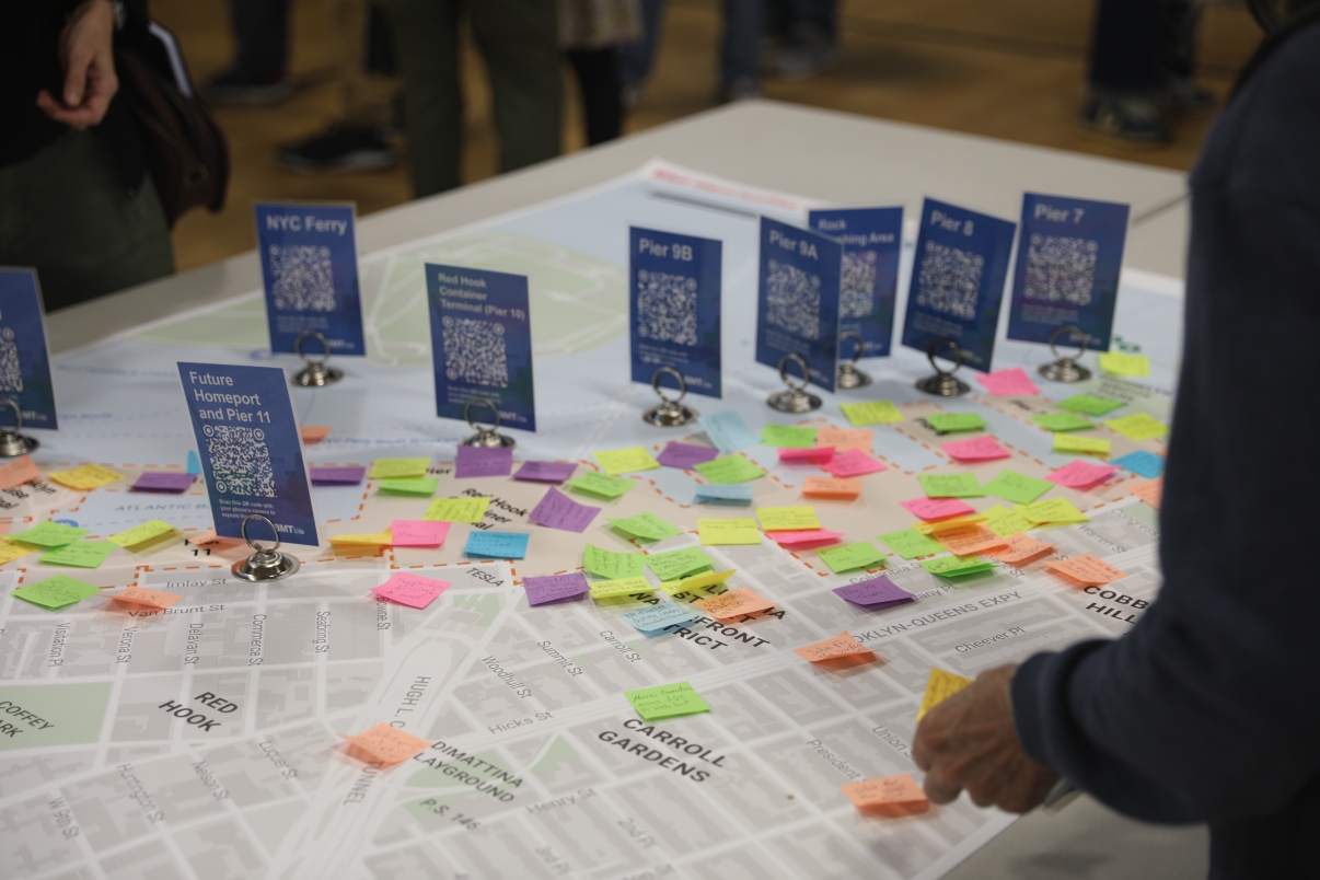 Person stands by a table with a large city map covered in colorful sticky notes and signs for Brooklyn Marine Terminal piers. Other people are blurred in the background.