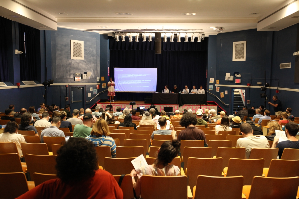 A group of people sit in an auditorium, facing a stage where several panelists are seated at a table. A person stands at a podium beside a projection screen displaying a presentation.