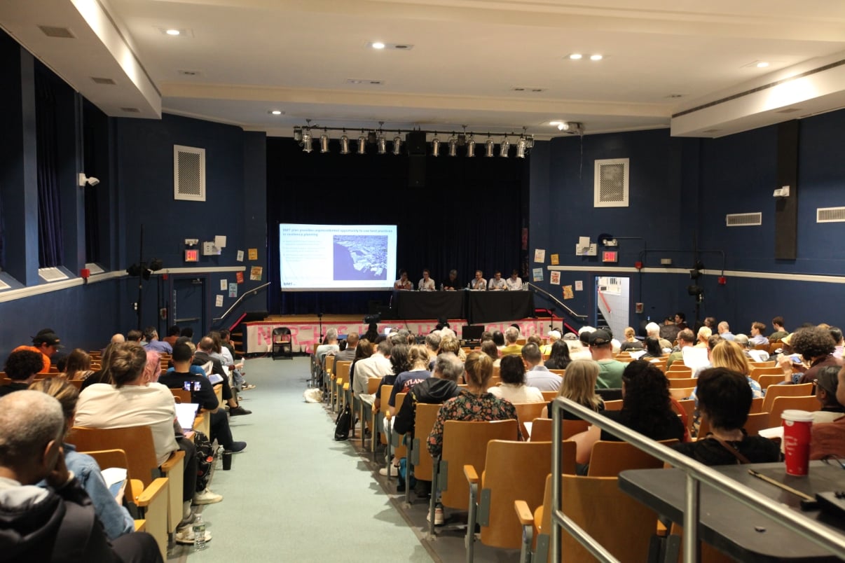 A diverse audience sits in rows of seats in an auditorium, watching a panel of speakers at a table on stage with a large projected presentation behind them.