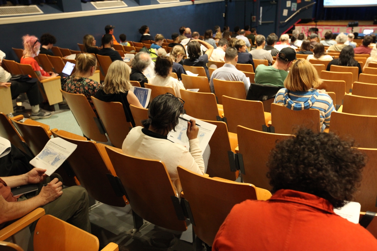 Audience members sit in rows of seats in a lecture hall, some reading papers, taking notes, or using laptops, while facing a presentation at the front of the room.