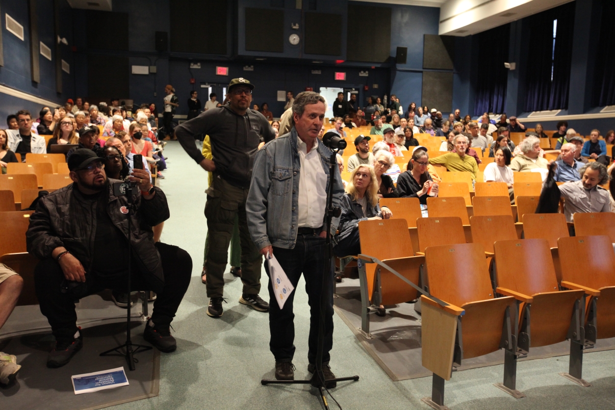 A man stands at a microphone in a crowded auditorium, speaking to an audience seated in rows of wooden chairs. Several people listen attentively, while others are engaged in their own conversations.