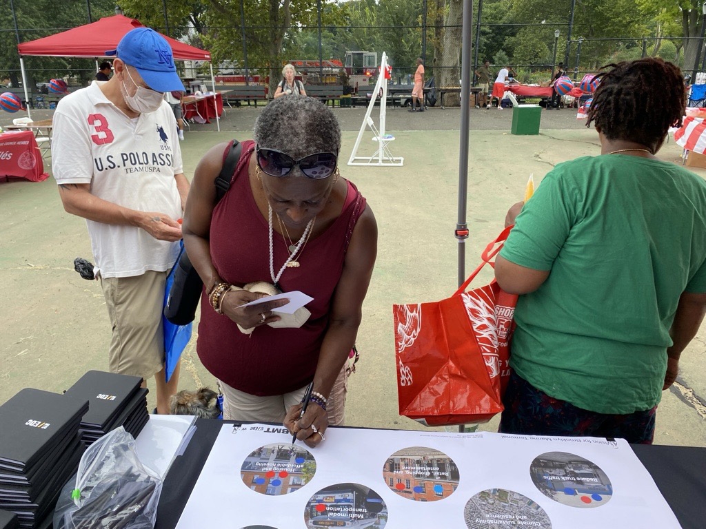 Three people stand at an outdoor table for NYCHA Family Day. A woman in a maroon top writes on a paper, a man in a white shirt stands nearby, and another person with a red bag faces away. There are papers and boxes on the table.