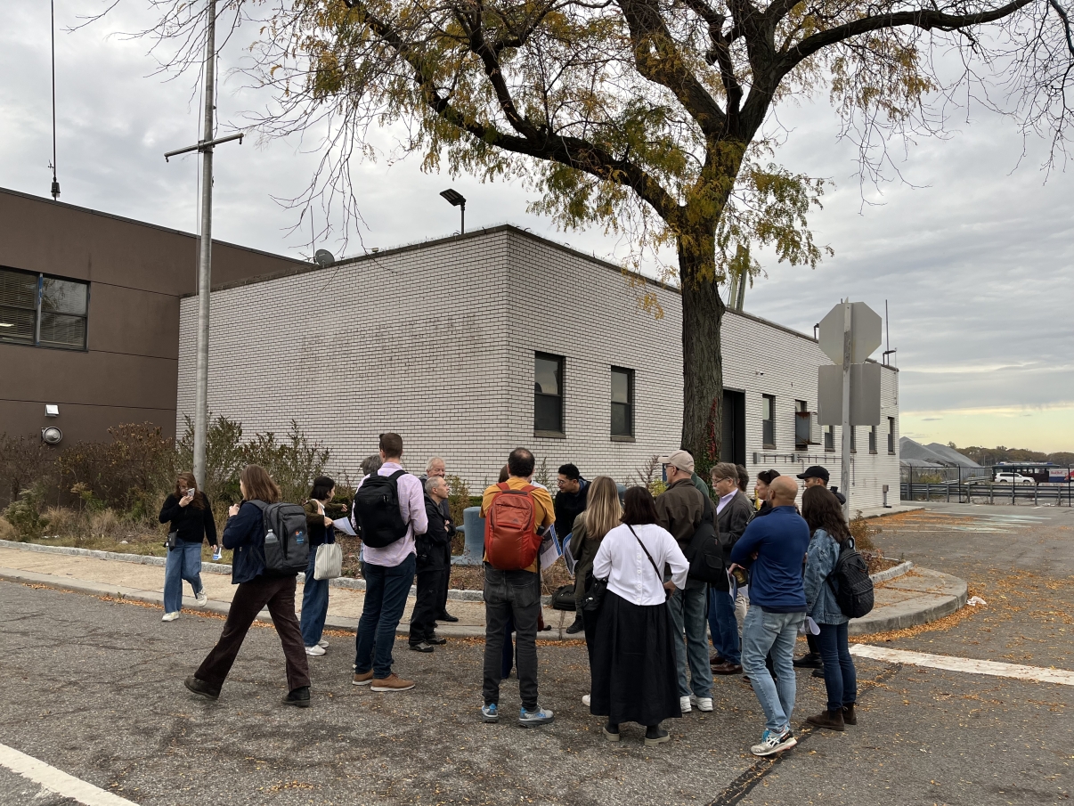 A group of people stands in a loose circle on a sidewalk near a white brick building and a tree with autumn leaves. They are there for the Brooklyn Marine Terminal Public Site Tour. Most are wearing jackets and backpacks, and the sky is overcast.