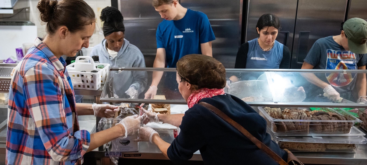 Six people stand behind a counter serving food in a cafeteria or soup kitchen, wearing gloves and focused on preparing and distributing meals. Various trays of food are visible in front of them. Six people stand behind a counter serving food in a cafeteria or soup kitchen, wearing gloves and focused on preparing and distributing meals. Various trays of food are visible in front of them.
