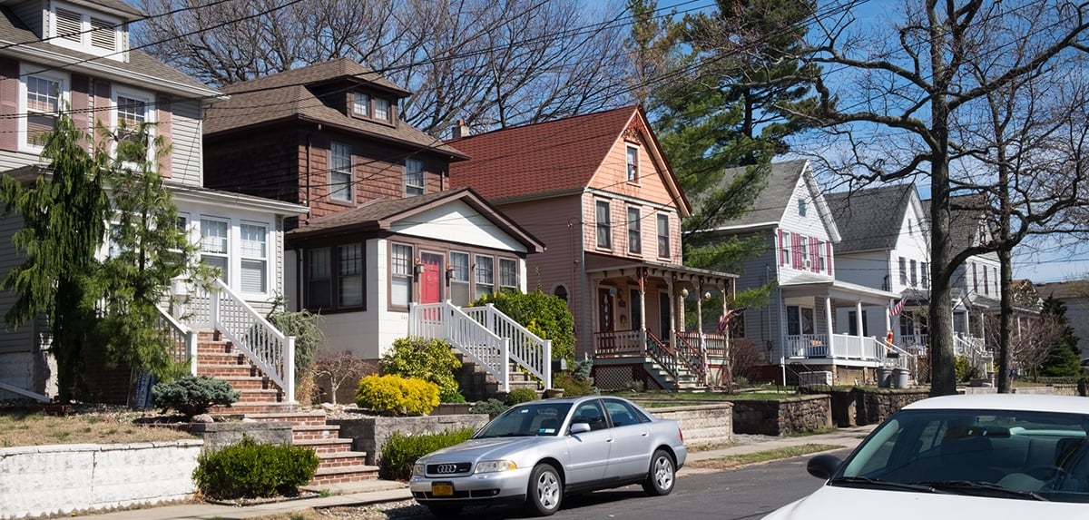 A row of colorful, two-story houses with porches and stairs along a suburban street. A silver car and a white car are parked on the street under leafless trees on a sunny day. A row of colorful, two-story houses with porches and stairs along a suburban street. A silver car and a white car are parked on the street under leafless trees on a sunny day.