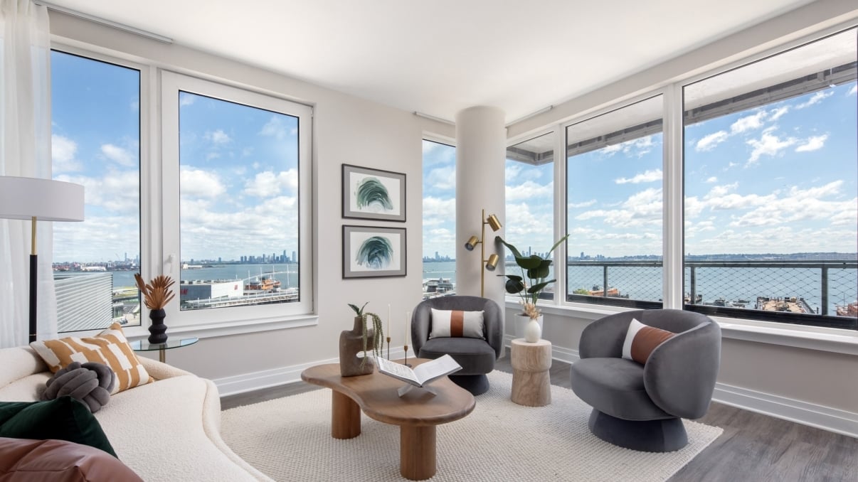 A modern, bright living room with large windows, gray armchairs, a white curved sofa, wooden coffee tables, plants, and a view of New York City skyline and water under a blue sky with clouds. A modern, bright living room with large windows, gray armchairs, a white curved sofa, wooden coffee tables, plants, and a view of New York City skyline and water under a blue sky with clouds.