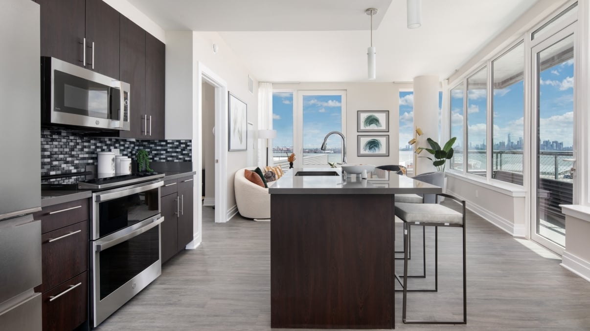 Modern kitchen with dark cabinets, stainless steel appliances, and a central island with two stools. Open living area with large windows showing a city skyline and waterfront view. Bright, minimalist decor with plants and artwork. Modern kitchen with dark cabinets, stainless steel appliances, and a central island with two stools. Open living area with large windows showing a city skyline and waterfront view. Bright, minimalist decor with plants and artwork.