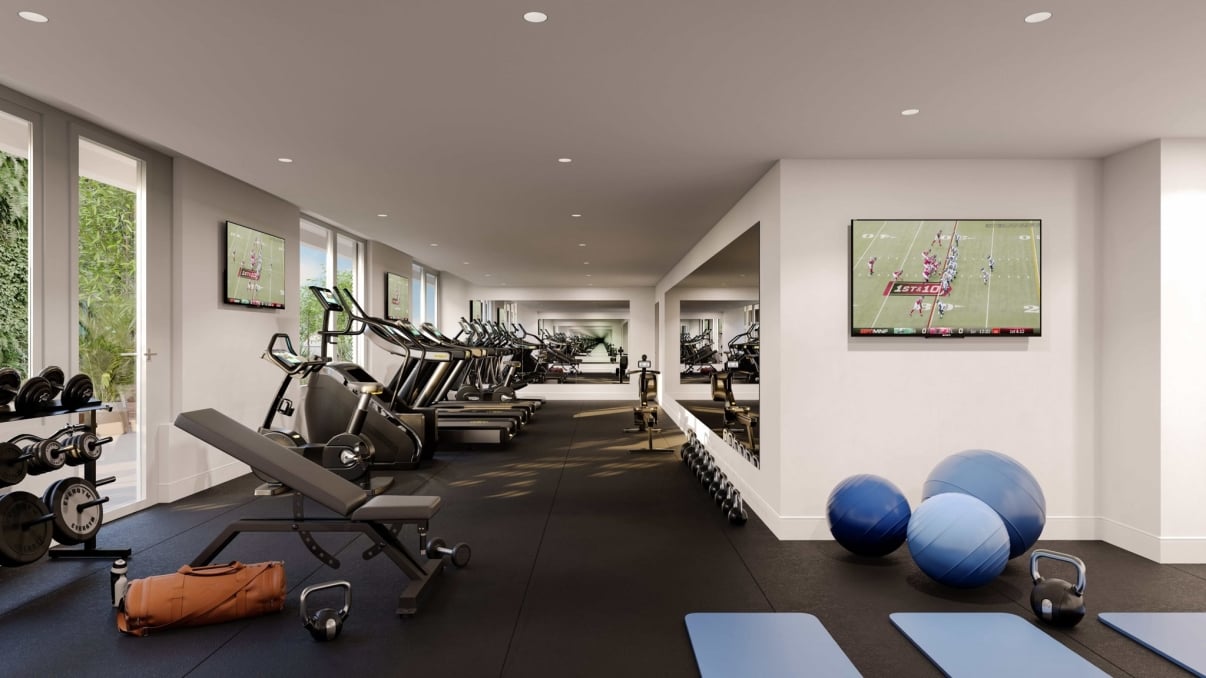 Modern gym with treadmills, weight benches, dumbbells, exercise balls, yoga mats, and large mirrors. Two TVs on the wall display a football game. Natural light enters through windows along the left side. Modern gym with treadmills, weight benches, dumbbells, exercise balls, yoga mats, and large mirrors. Two TVs on the wall display a football game. Natural light enters through windows along the left side.