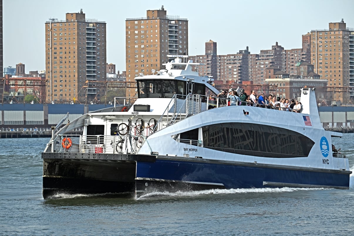 A NYC ferry with passengers on the upper deck travels on the water, with city buildings and apartment complexes visible in the background on a clear day.