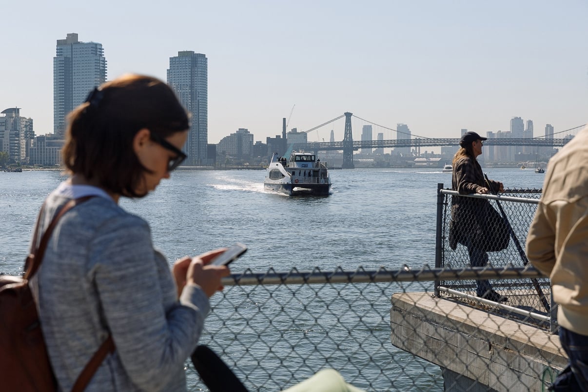 A woman using her phone sits by a fence overlooking a river, with a NYC Ferry approaching and city buildings and a bridge visible in the background under a clear sky.