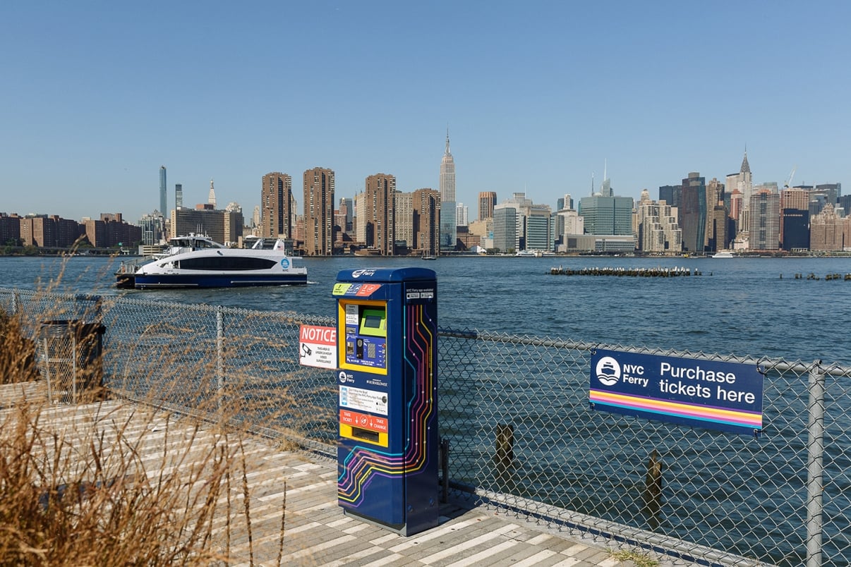A NYC Ferry ticket kiosk on a pier overlooks the East River with an NYC Ferry passing by and the Manhattan skyline, including the Empire State Building, visible in the background.