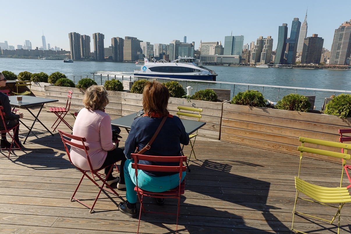 Two people sit at a table on a wooden outdoor deck with colorful chairs, overlooking a river with a NYC Ferry boat passing by and a city skyline in the background on a sunny day.