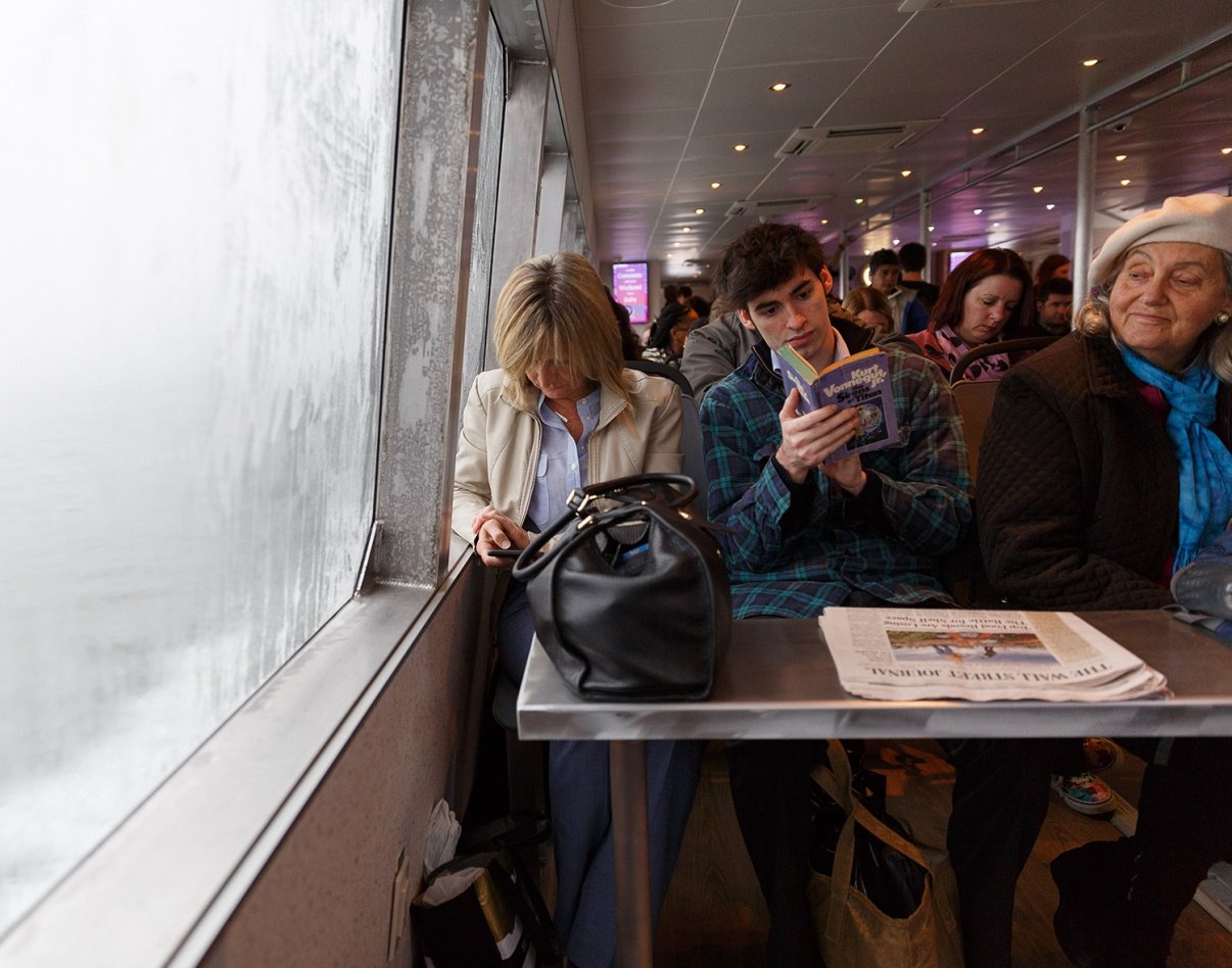 People sitting closely together on a NYC Ferry; a woman looks down at her phone, a young man reads a guidebook, and an elderly woman in a white beret gazes forward. A newspaper and a black bag are on the table.
