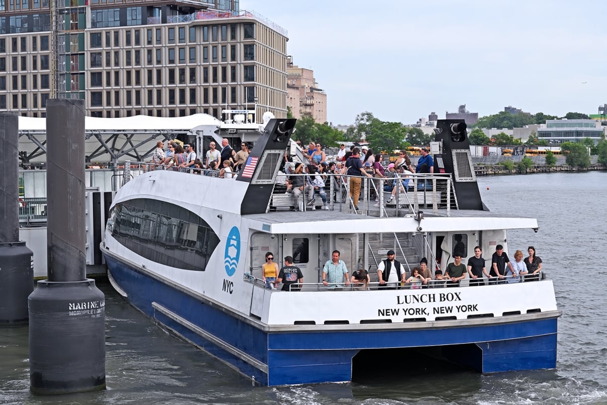 A crowded NYC Ferry named LUNCH BOX with passengers on the upper and lower decks arrives at a dock in New York City on a sunny day; buildings and greenery are visible in the background.