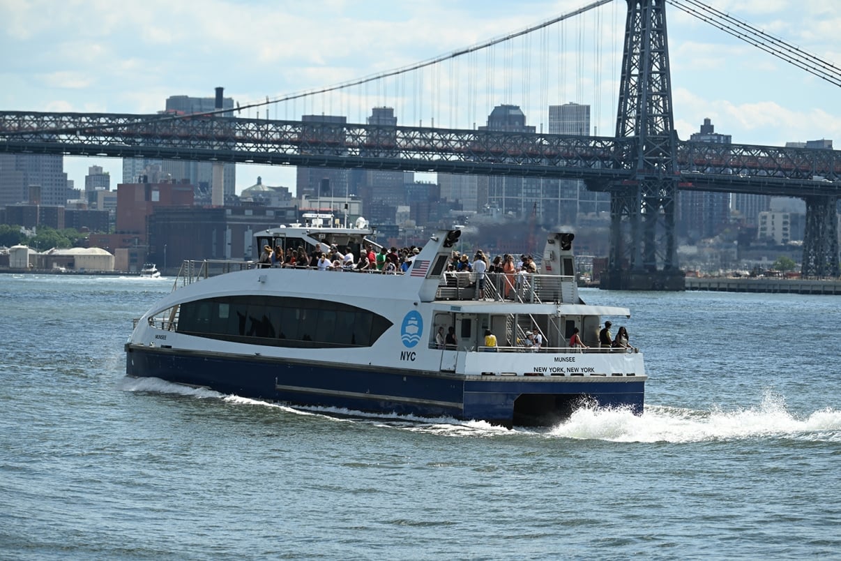 A NYC Ferry carrying passengers travels on the water with a city skyline and a large bridge in the background on a clear day.