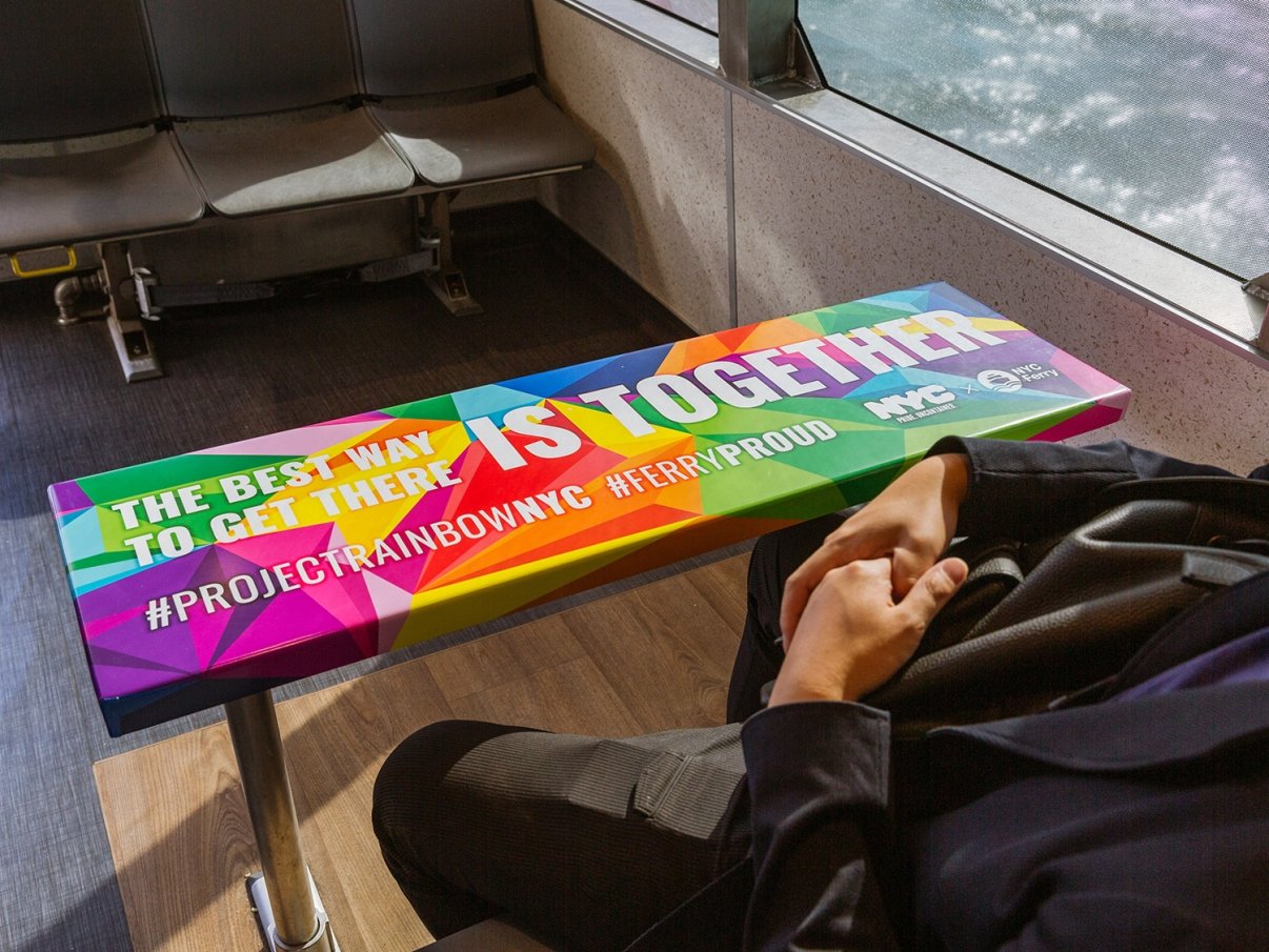 A person sits on a NYC Ferry next to a colorful table with the text The best way to get there is together and hashtags #PROJECTRAINBOWNYC and #FERRYPROUD, with the NYC logo.