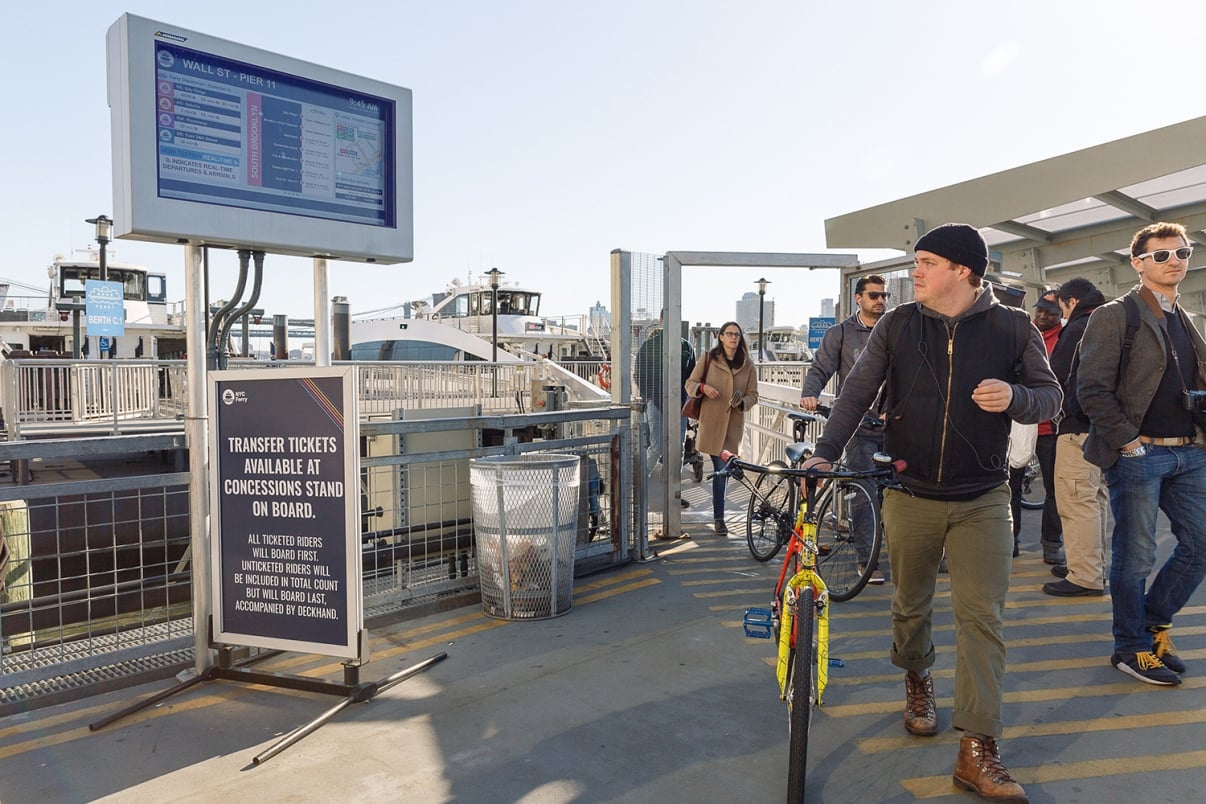 A man walks a bicycle past a group of people at the Wall Street NYC Ferry terminal, with boats docked in the background and a large electronic schedule board overhead.