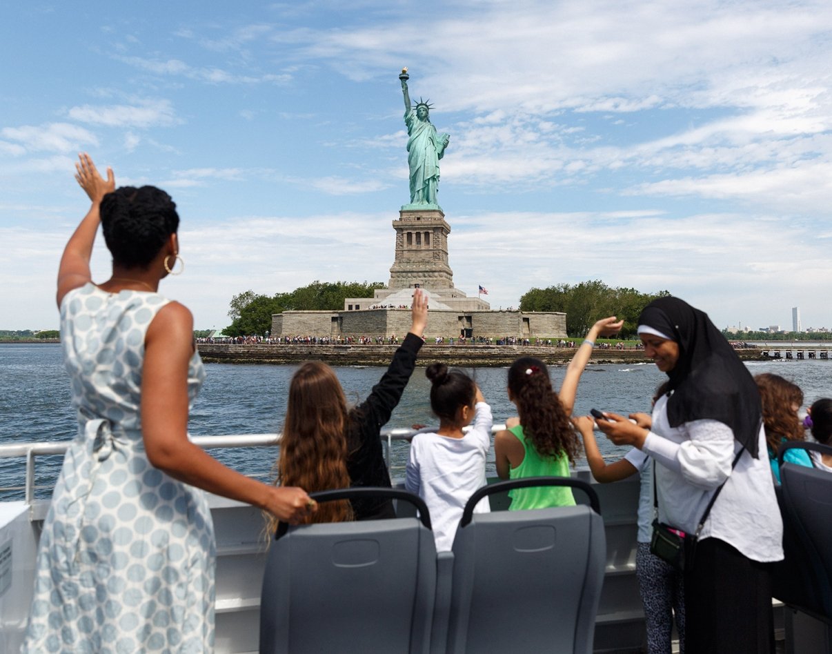 People on a NYC Ferry wave to the Statue of Liberty on Liberty Island under a partly cloudy sky, with some taking photos and others looking at the monument.