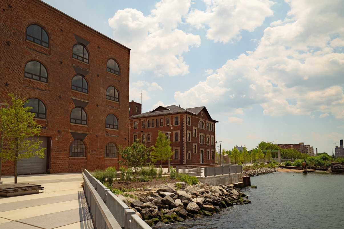 Red brick industrial buildings sit beside a river, surrounded by trees and rocks along the water’s edge. The sky is partly cloudy, and the area appears calm and well-maintained.
