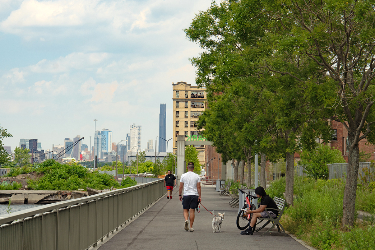 A man walks a dog on a riverside path lined with trees; another man jogs, and a third sits on a bench by a bicycle. City skyline and buildings are visible in the background under a partly cloudy sky.
