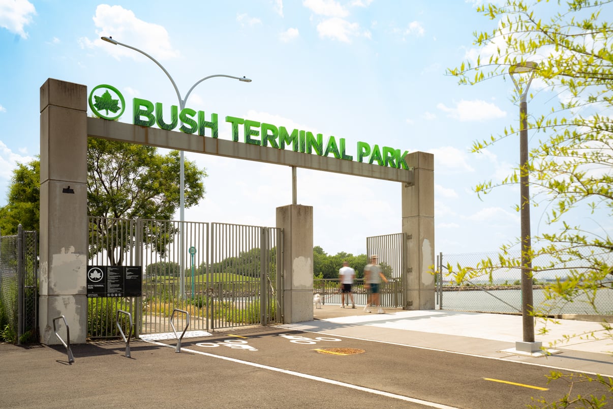 Entrance gate to Bush Terminal Park with green sign, wide open doors, trees, fencing, and a few blurred people walking inside under a bright blue sky with scattered clouds.