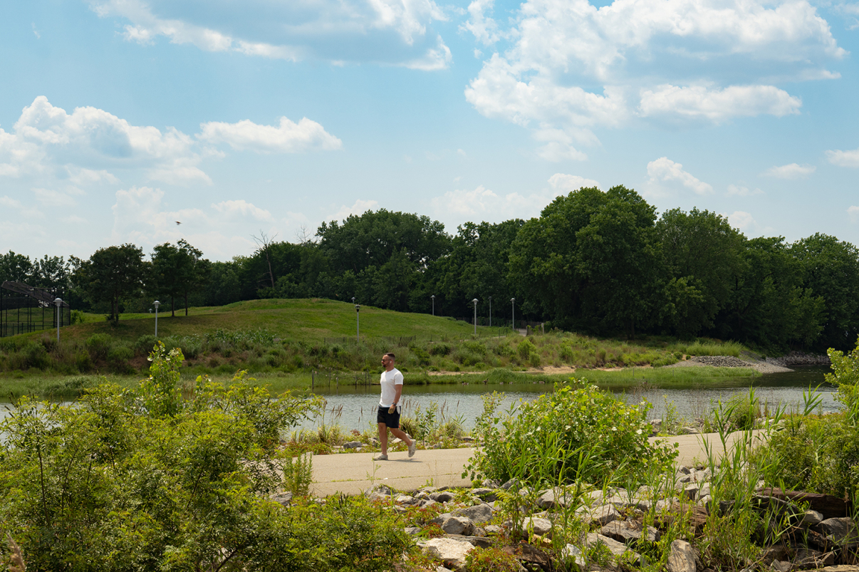 A man in a white shirt and black shorts walks on a paved path beside a river, surrounded by green bushes and trees under a partly cloudy sky.